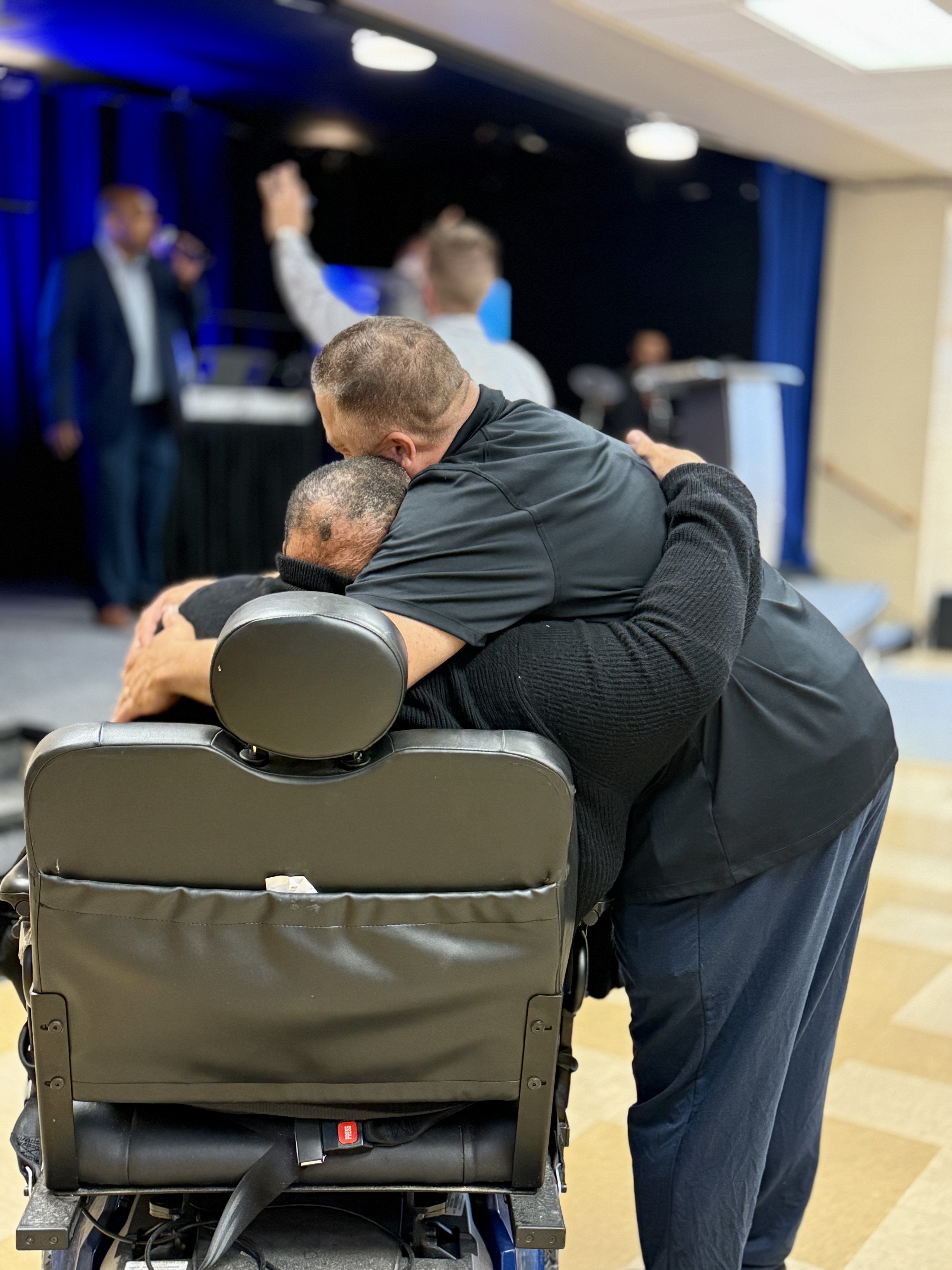 Men embracing in powerful prayer moment with wheelchair at Wisconsin Men's Ministry gathering